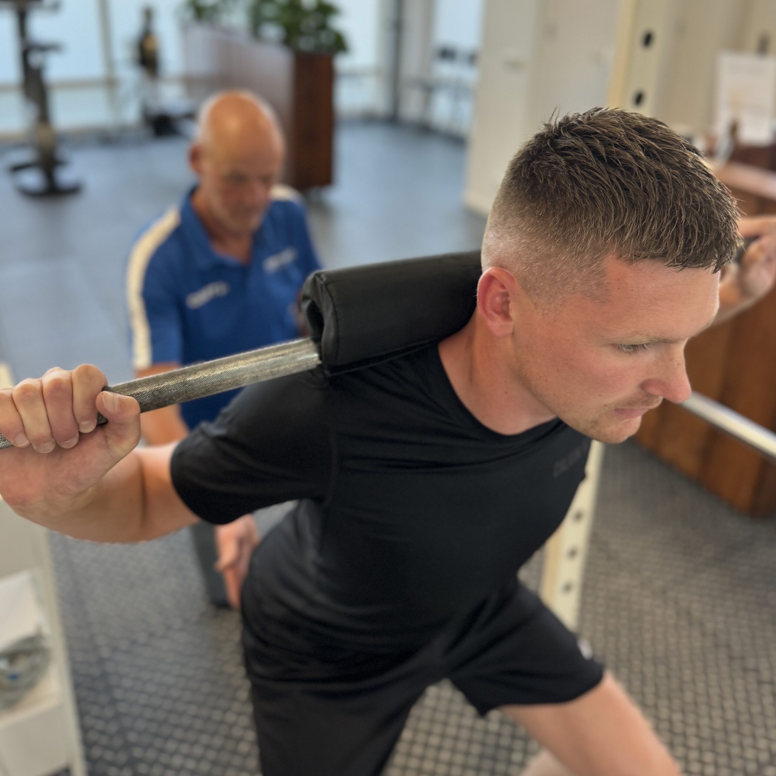 Fysiotherapeut begeleidt patiënt bij squat met halter tijdens revalidatie na een achillespeesruptuur